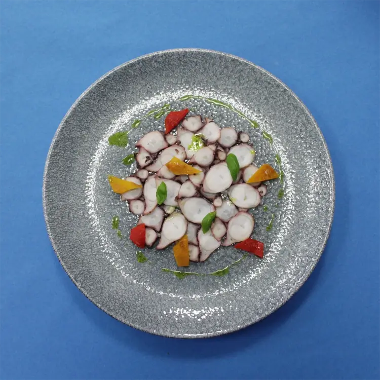 Estetycznie podane carpaccio z ośmiornicy z warzywami i oliwą ziołową – nowoczesna przystawka w stylu fine dining, zaprezentowana na talerzu ceramicznym na niebieskim tle.