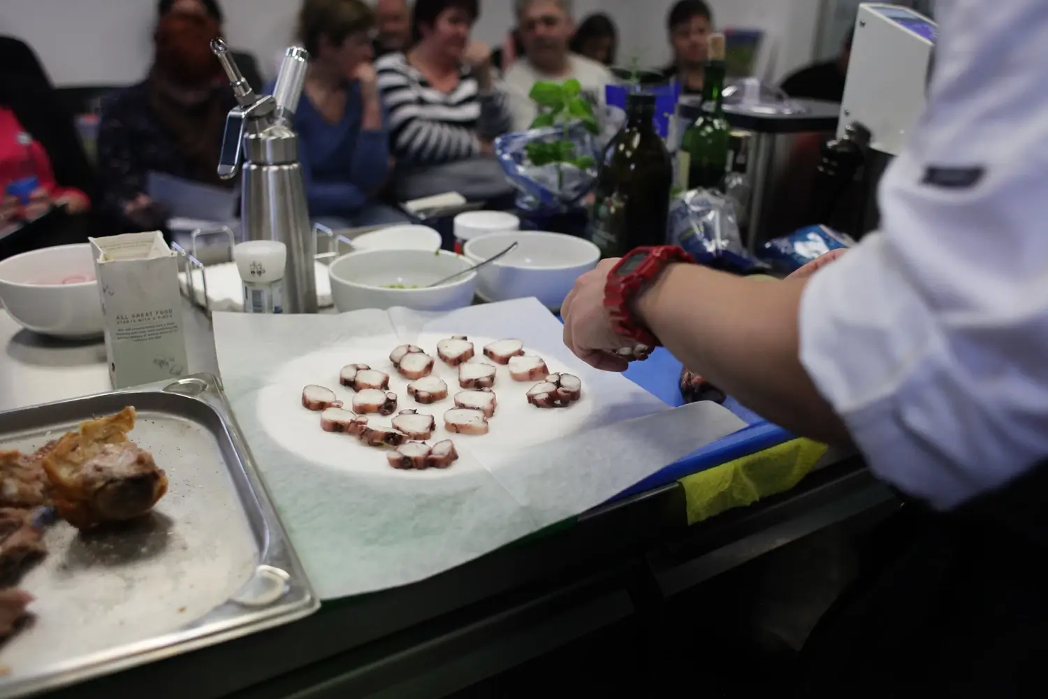 Przygotowanie carpaccio z ośmiornicy podczas pokazu kulinarnego – kucharz układa plasterki ośmiornicy na pergaminie, w tle widoczna publiczność i stanowisko z produktami.