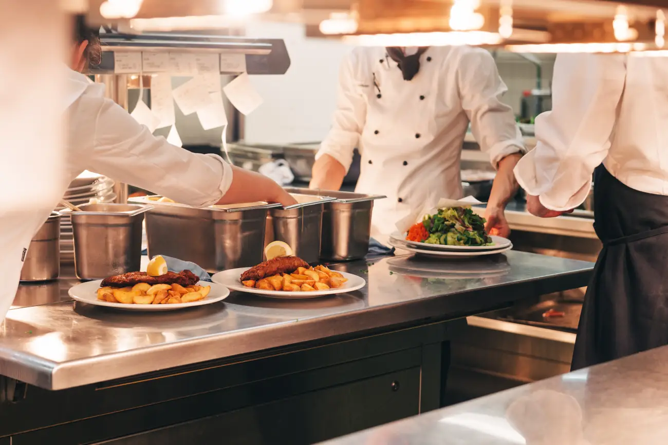 Pracownicy kuchni w restauracji przygotowujący potrawy przy stanowiskach z garnkami i patelniami, w tle profesjonalny piec i okap.