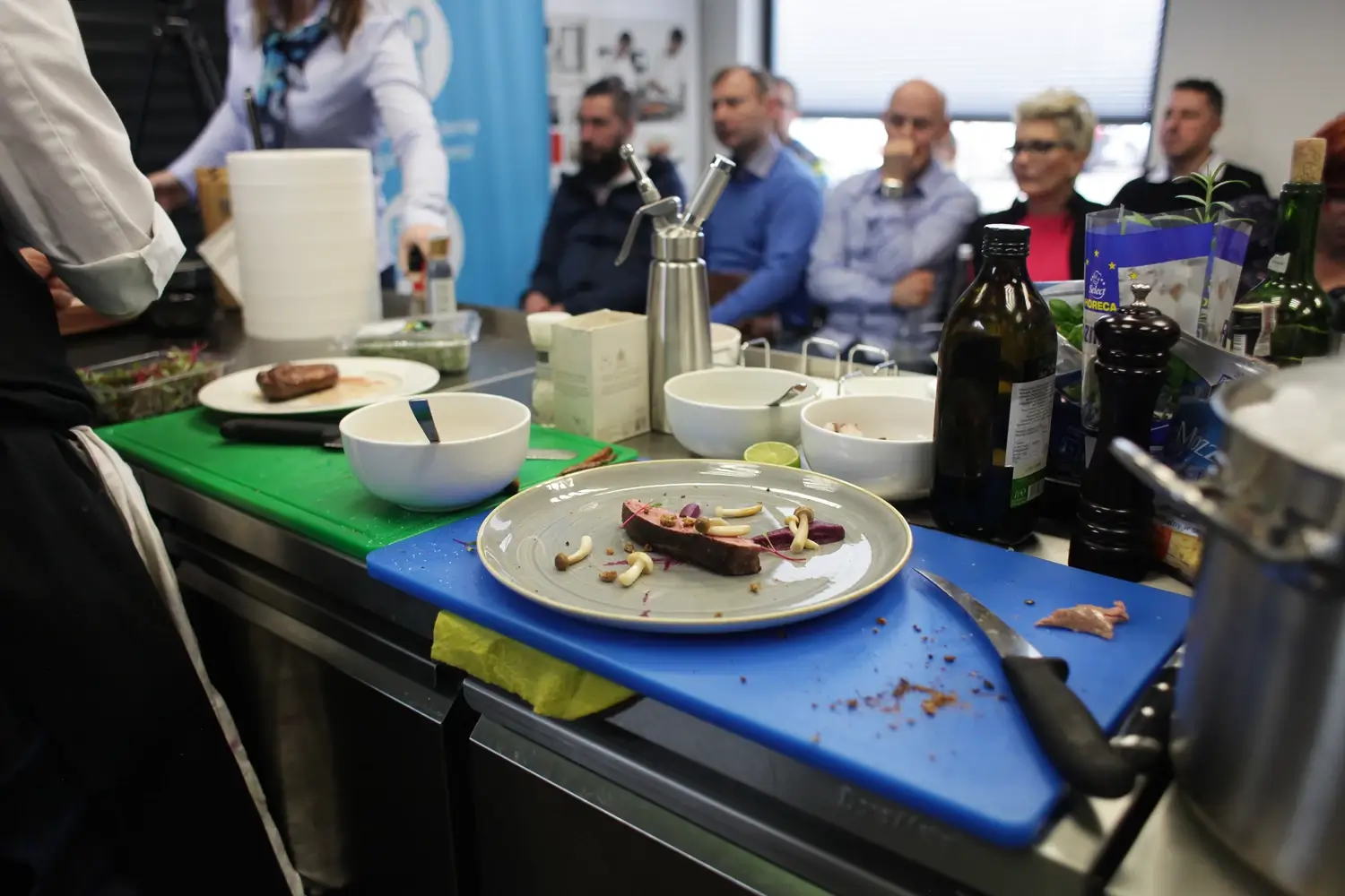 Pokaz kulinarny na żywo – przygotowanie piersi z kaczki na purée z kapusty w obecności publiczności, z widocznymi deskami do krojenia, półproduktami i narzędziami kuchennymi na blacie roboczym.