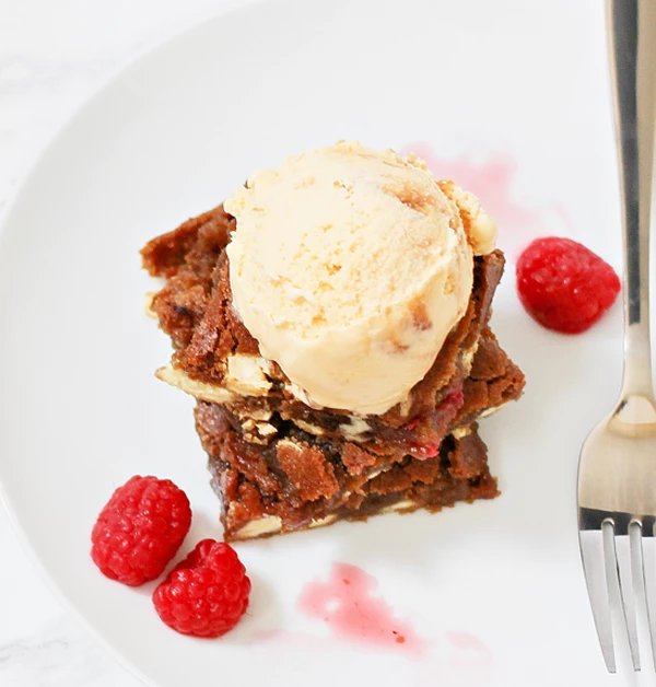 Porcja brownie z lodami waniliowymi, podana na białym talerzu z malinami i odrobiną sosu owocowego, obok widelczyk deserowy.
