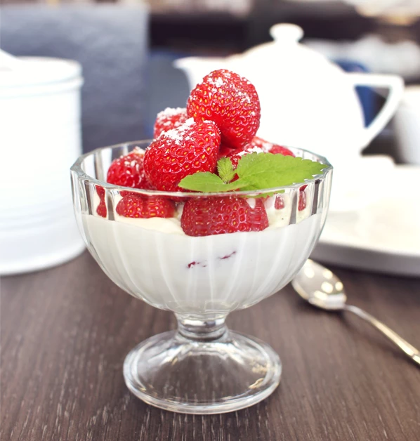 Deser z musu mascarpone podany w eleganckim szklanym pucharku, udekorowany świeżymi truskawkami, listkami mięty i oprószony cukrem pudrem.