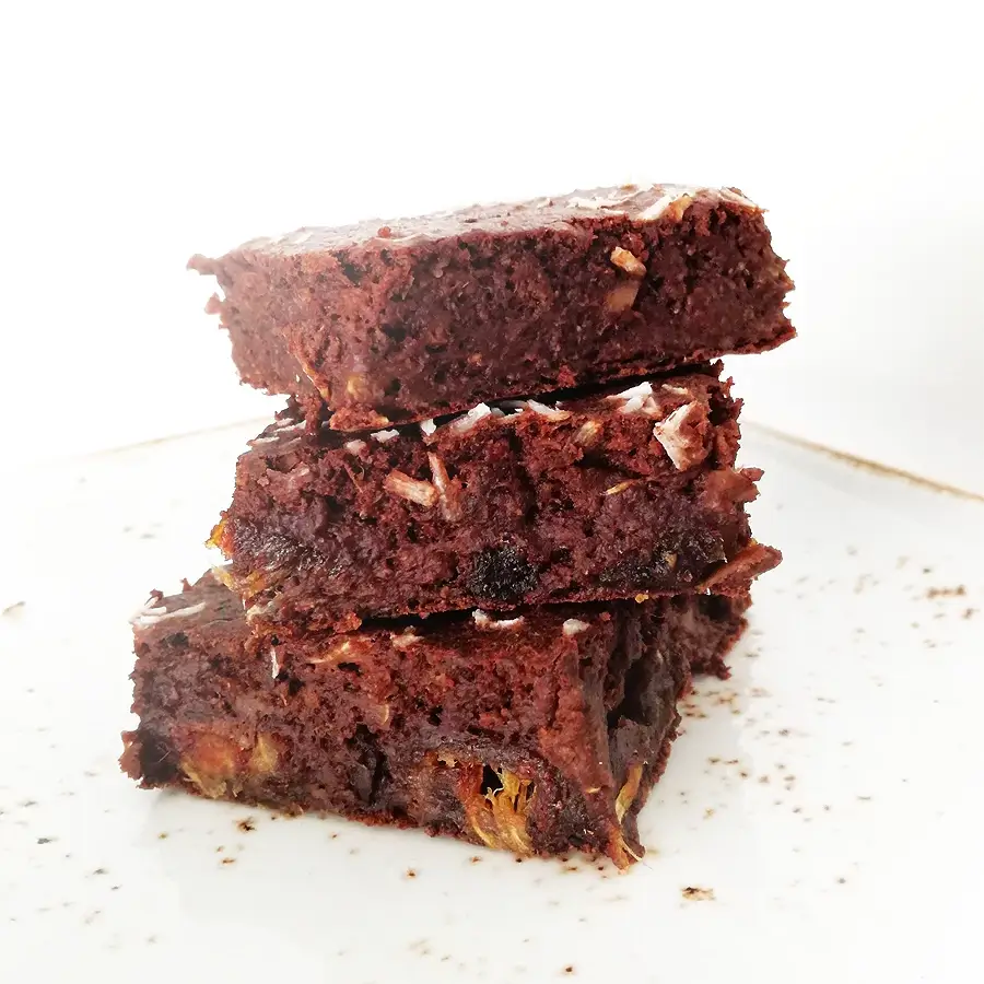 Trzy kawałki czekoladowego brownie z bakaliami, ułożone jeden na drugim na białym talerzu.