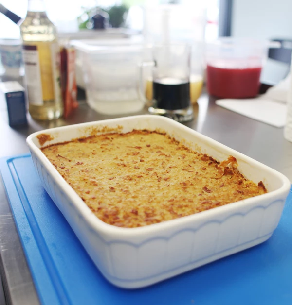 Babka ziemniaczana upieczona na złoty kolor, podana w białym ceramicznym naczyniu żaroodpornym, ustawionym na niebieskiej desce, w tle kuchenne akcesoria i butelki z przyprawami.
