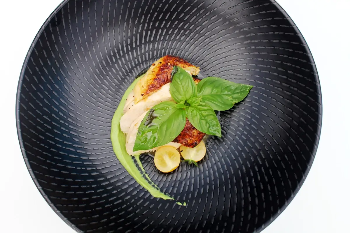 Elegancko podany filet z kurczaka przygotowany metodą sous vide, ułożony na czarnym talerzu z dodatkiem zielonych ziół i żółtych warzyw, ozdobiony listkiem bazylii i dekoracyjnym sosem.