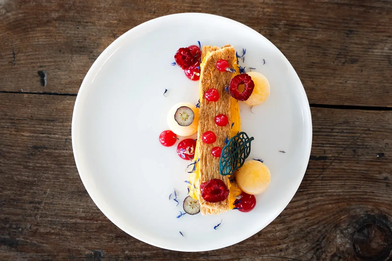 Deser mille-feuille z kremem kokosowo-dyniowym, udekorowany świeżą pomarańczą, kulkami musu oraz owocami dzikiej róży, podany na białym talerzu na tle drewnianego stołu. Kompozycja artystyczna z dodatkiem jadalnych kwiatów i ozdoby z masy cukrowej w kształcie liścia.