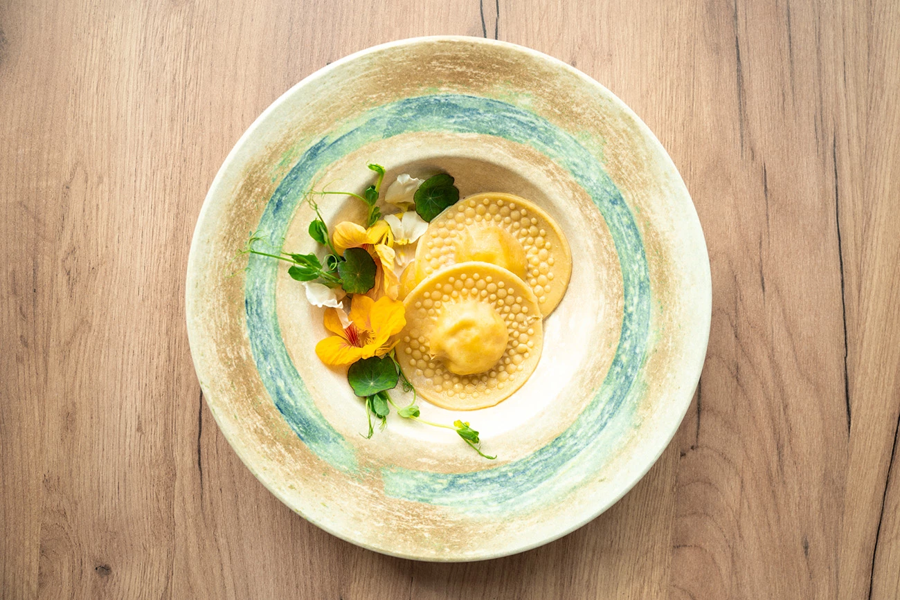 Ravioli z konfiturą pomarańczową i kremem kawowym z migdałami, elegancko ułożone na ozdobnym porcelanowym talerzu. Danie udekorowane świeżymi listkami groszku i kolorowymi jadalnymi kwiatami, serwowane na tle jasnego, drewnianego blatu.