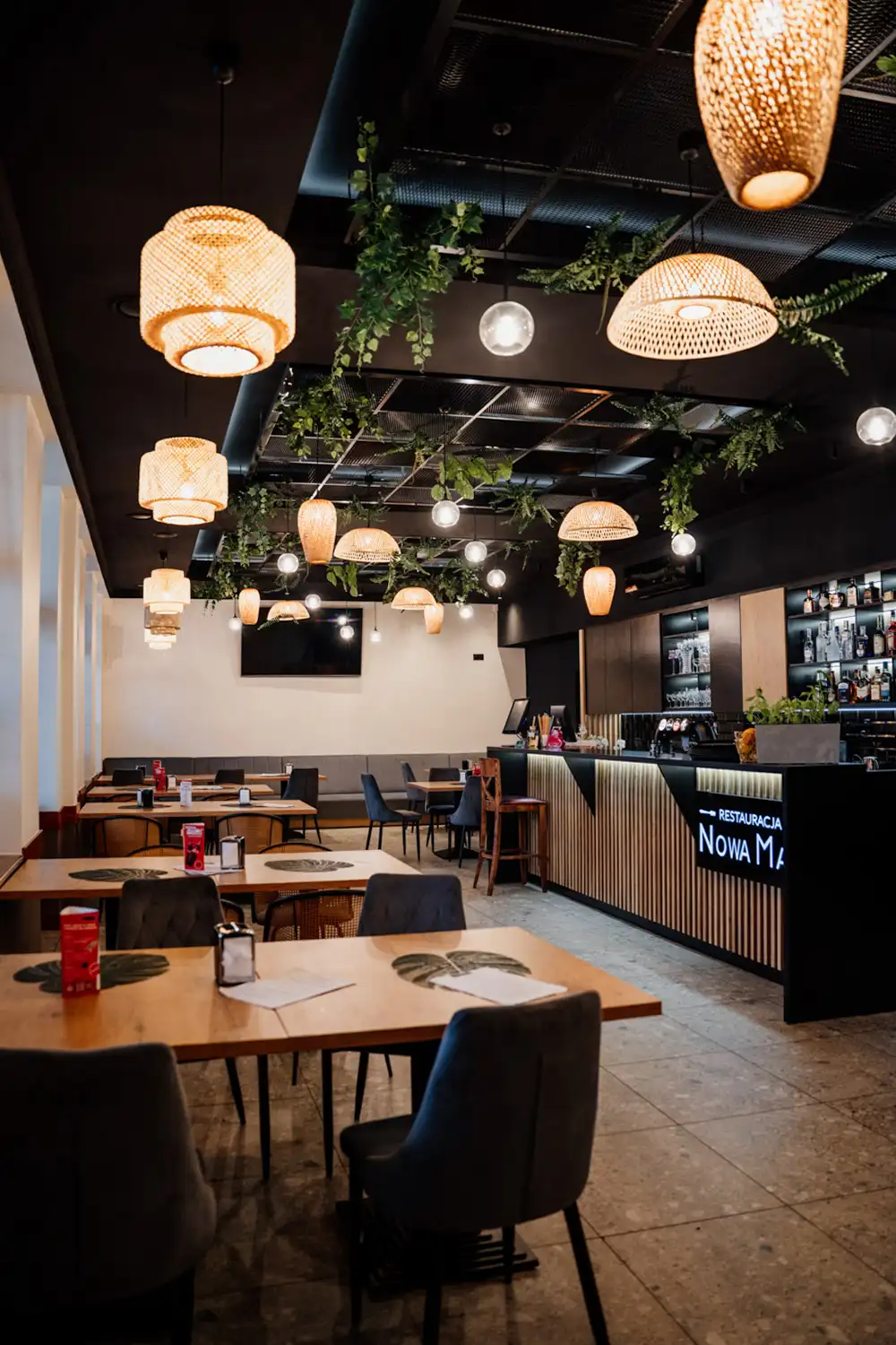 Sala restauracyjna Nowa Maja w Skoczowie z drewnianymi stołami, tapicerowanymi krzesłami, barem oraz dekoracyjnym oświetleniem i zielenią sufitową.