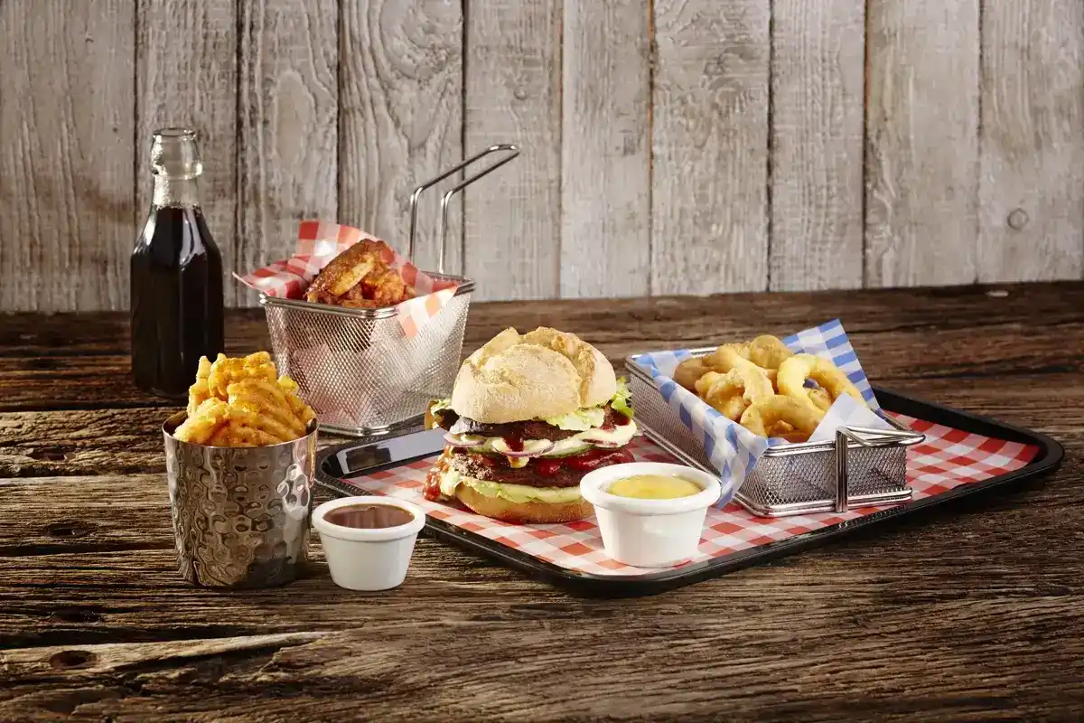 Kompletne danie fast food na tacy w stylu retro, składające się z burgera z warstwą mięsa, warzyw i sosu, podanego z frytkami w metalowej misce, porwowanymi krążkami cebulowymi w koszyczku oraz frytkami w kratkę. Obok znajduje się butelka napoju gazowanego i sos w małym pojemniku. Całość podana na tacy wyłożonej czerwoną i białą kratką, z drewnianym tłem, które nadaje rustykalny charakter.