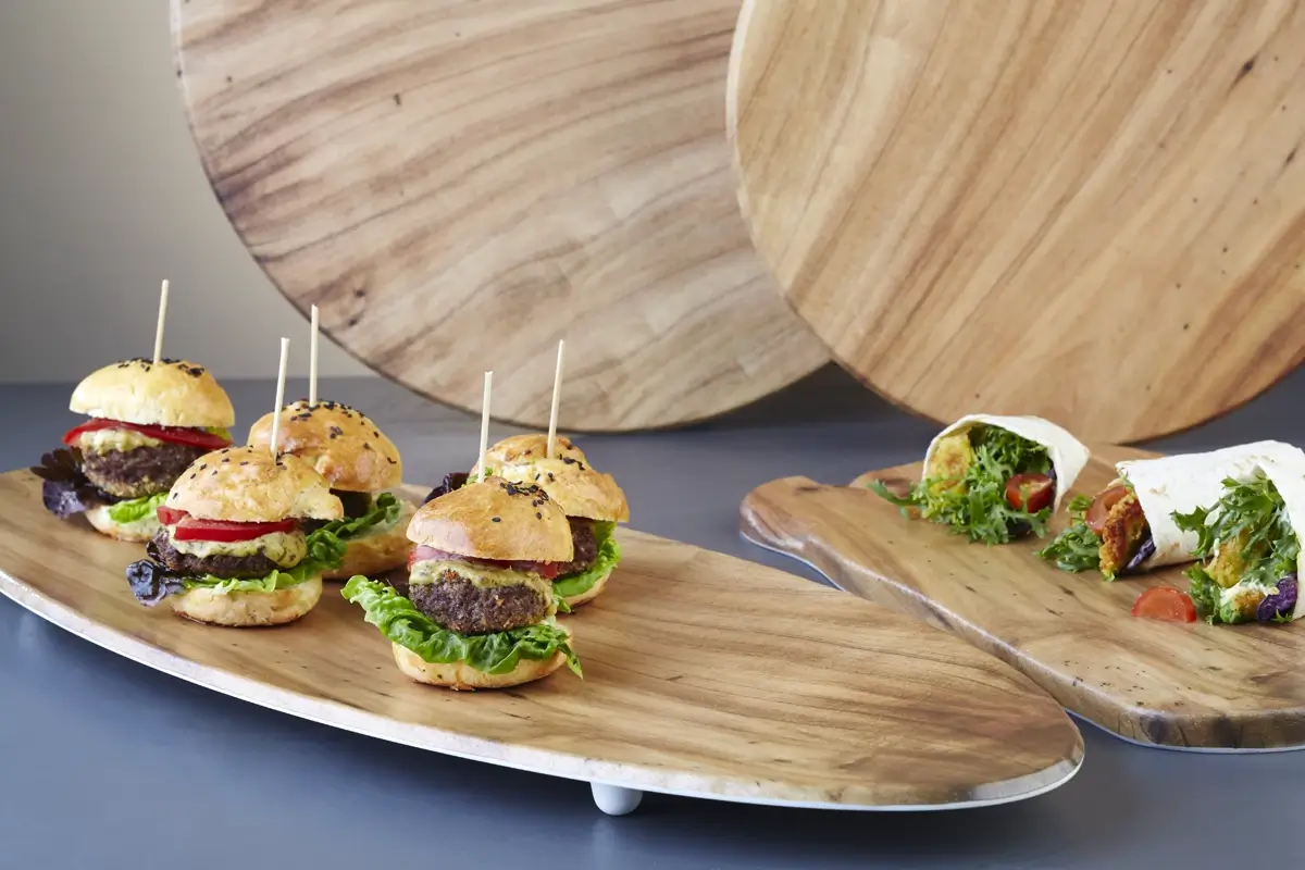 Kanapki z burgerami i świeżymi warzywami na drewnianej desce, obok zawijane tortille z warzywami. Tło stanowią drewniane okrągłe deski.