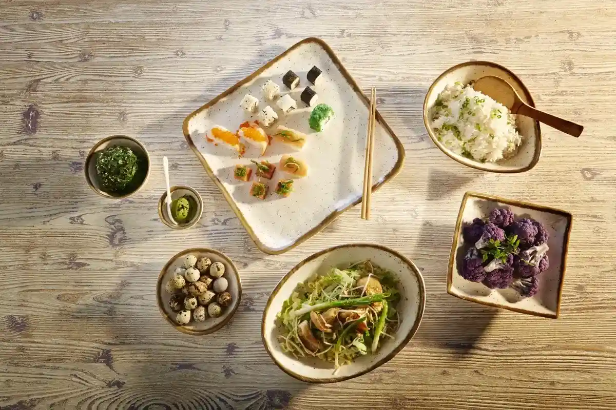 Zestaw azjatyckich potraw na różnych talerzach i miseczkach: sushi z wodorostami i wasabi, gotowany ryż z cebulką, danie z pieczonymi jajkami przepiórczymi, sałatka z warzyw i smażony kalafior w misce.