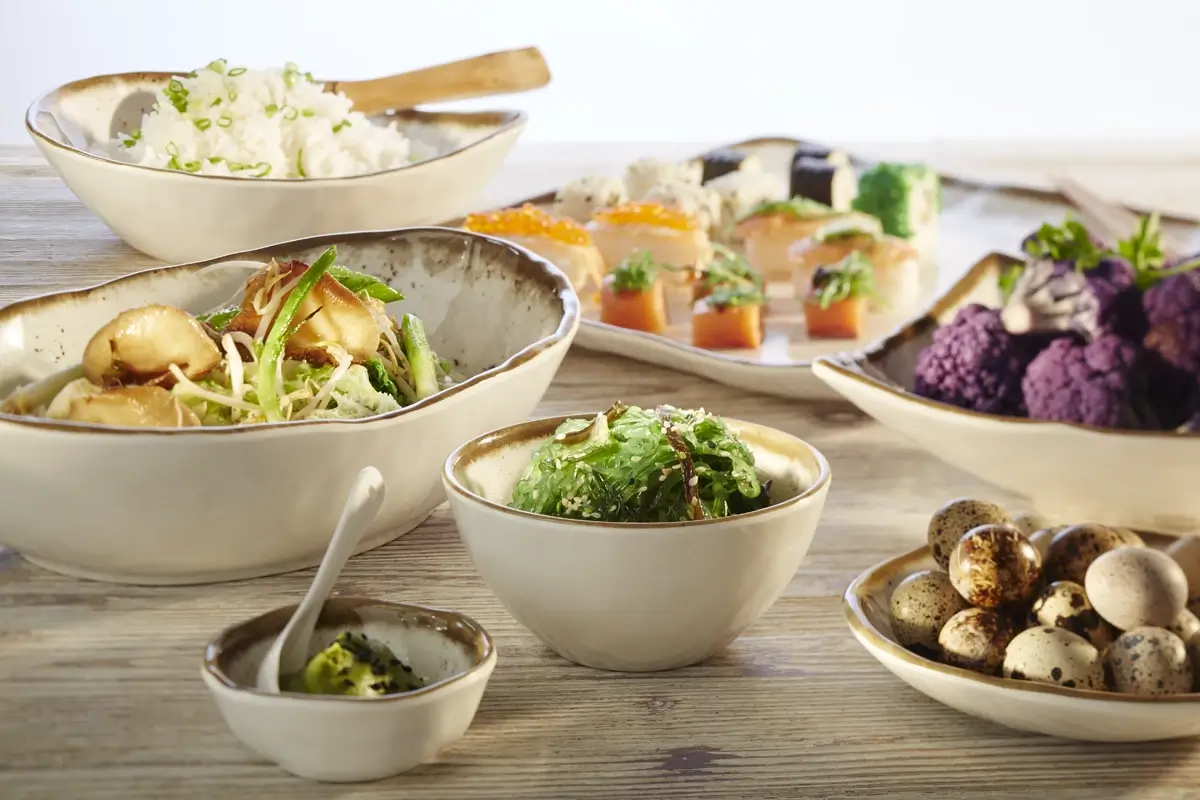 Azjatycki posiłek składający się z różnych potraw: sushi, gotowanego ryżu z cebulką, sałatki z warzywami, wodorostów i jajek przepiórczych, podane w miseczkach i talerzach w ciepłych odcieniach beżu.