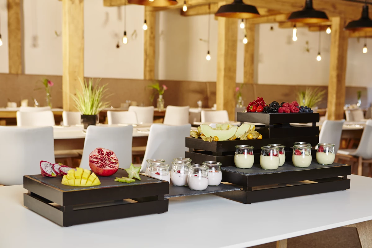 Aranżacja stołu bufetowego z czarnymi drewnianymi skrzynkami zawierającymi wybór świeżych owoców, w tym granat, mango i smoczy owoc, a także słoiki z jogurtem z jagodami na wierzchu. Aranżacja jest starannie zorganizowana z różnymi warstwami skrzynek, prezentując kolorową i zdrową ofertę. W tle znajdują się stoły jadalne ustawione w nowoczesnej restauracji z delikatnym oświetleniem i drewnianymi akcentami, tworząc zapraszającą i przytulną atmosferę.