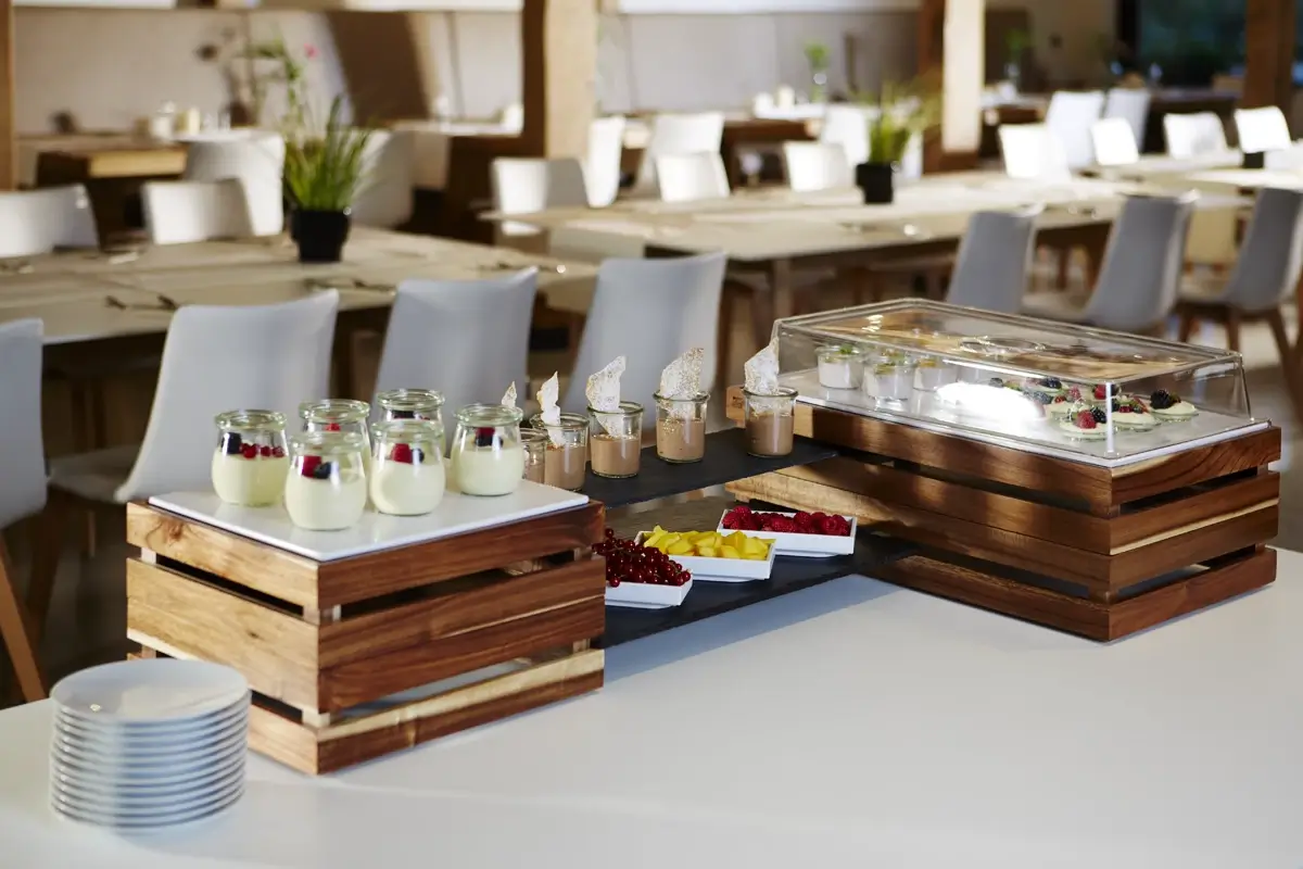 Deserowy bufet w eleganckiej restauracji. Na drewnianych skrzynkach umieszczono słoiczki z deserami (jogurt z owocami) oraz kremowe deserki w szklanych pojemnikach. W tle widać świeże owoce: maliny, mango i granat. Na stole także plastikowa osłona z ciastkami oraz talerze czekające na gości.