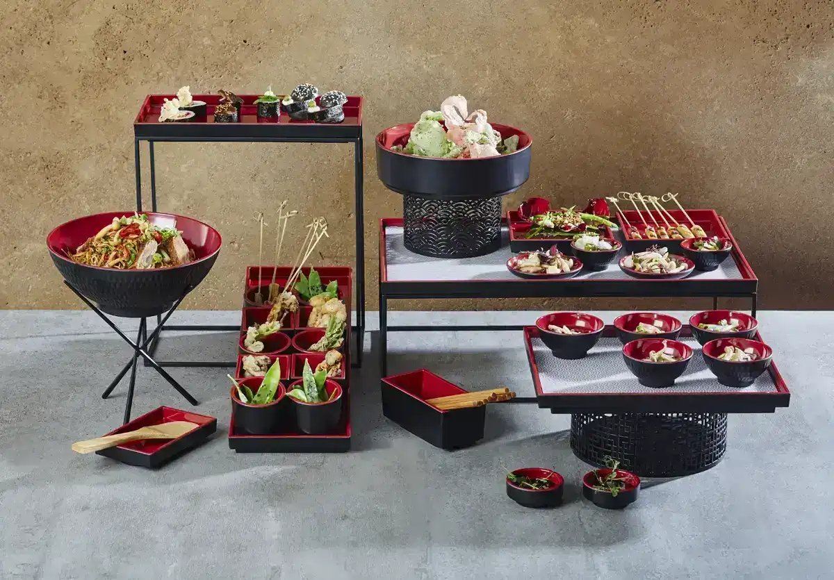 Stylowa prezentacja azjatyckich dań na kilku poziomach eleganckiego serwisu. Na pierwszym planie znajdują się czerwono-czarne miski z różnymi potrawami, w tym sushi, sałatkami oraz przekąskami, jak krewetki i warzywa. W tle duża miska z makaronem i dużym pojemnikiem na lody. Całość uzupełniają drewniane pałeczki i szpikulce do serwowania, a kompozycja jest ustawiona na industrialnym tle w odcieniach brązu i szarości.