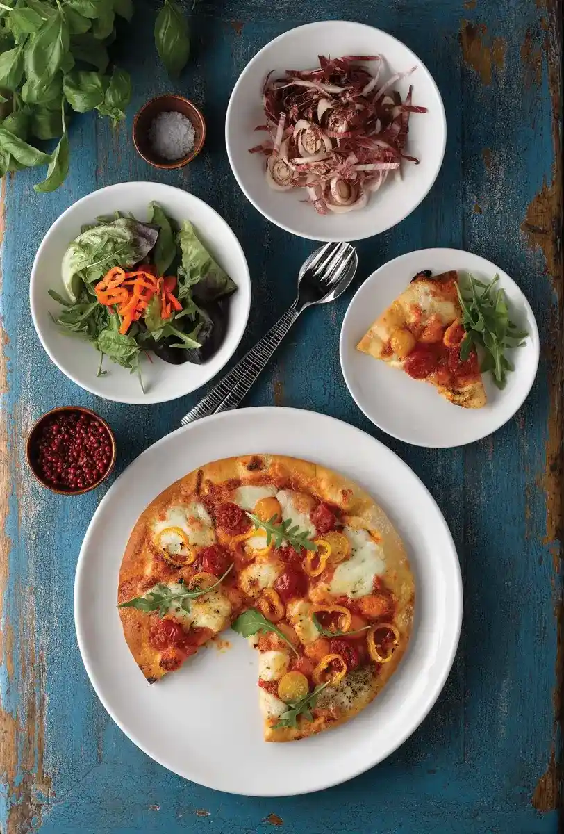 Pizza z pomidorami i rukolą podana na białym talerzu, obok miseczki z kolorowymi sałatami, talerzyka z kawałkiem pizzy i rukolą oraz dodatkami w miseczkach, na niebieskim, postarzanym stole.