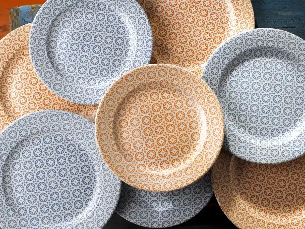 Sześć ceramicznych talerzy z geometrycznym wzorem w stylu retro, w dwóch kolorach – niebieskim i pomarańczowym, ułożone częściowo jeden na drugim na niebieskim drewnianym stole.