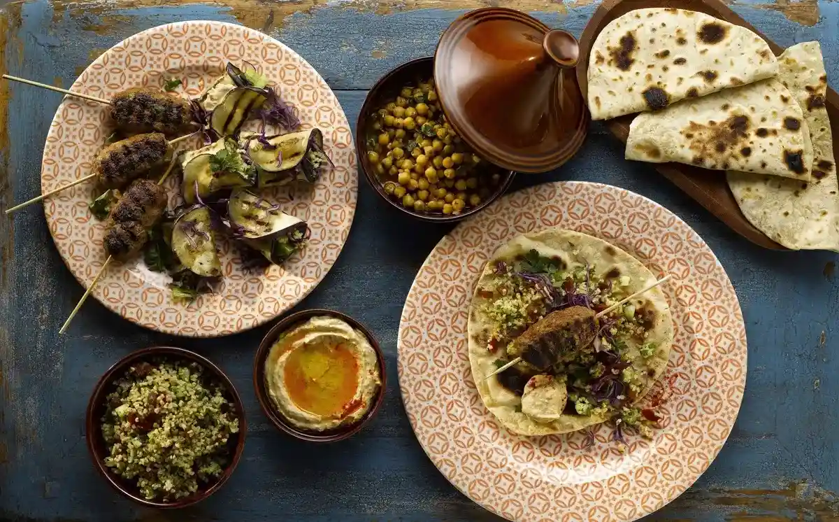 Kompozycja kuchni bliskowschodniej – talerze z grillowanymi szaszłykami, pieczonymi warzywami i lawaszem, miseczka hummusu, sałatka tabbouleh, ciecierzyca w glinianym naczyniu typu tagine oraz kawałki świeżego płaskiego chleba, wszystko ułożone na niebieskim, drewnianym stole.