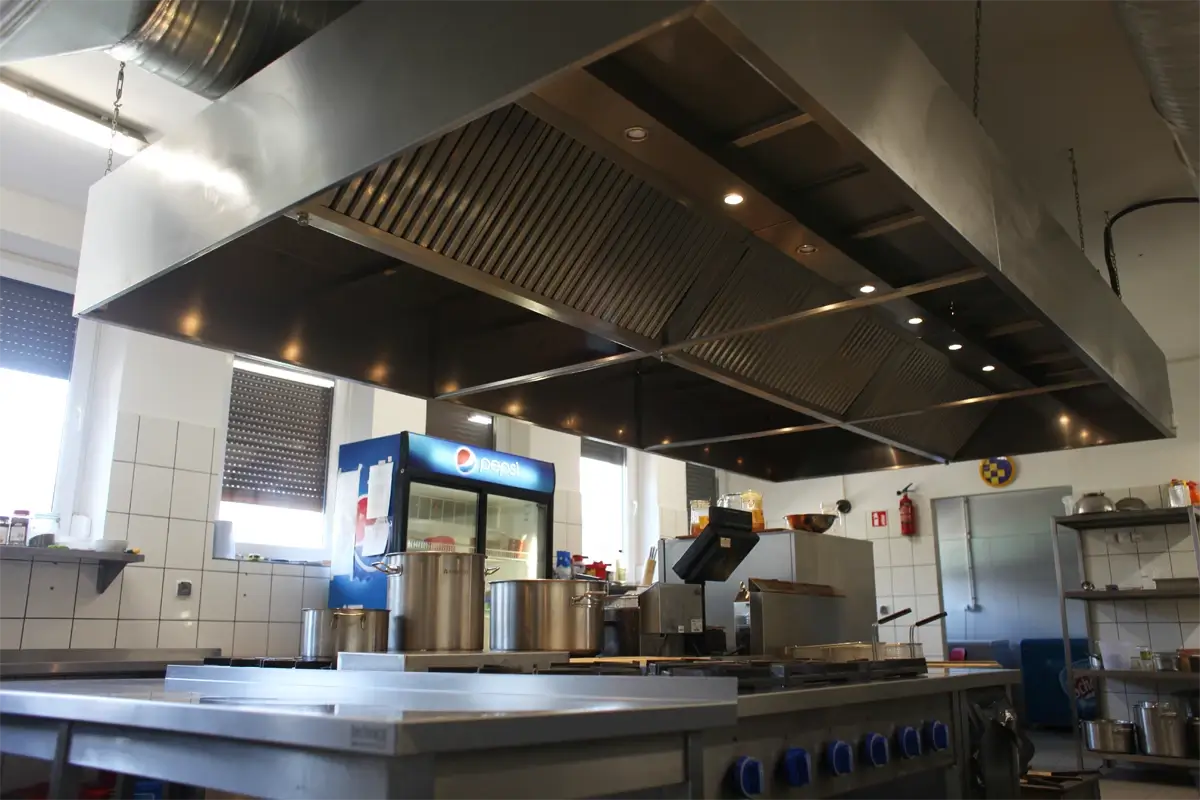Centralny okap wentylacyjny w kuchni gastronomicznej nad kuchnią gazową i wyspą roboczą.
