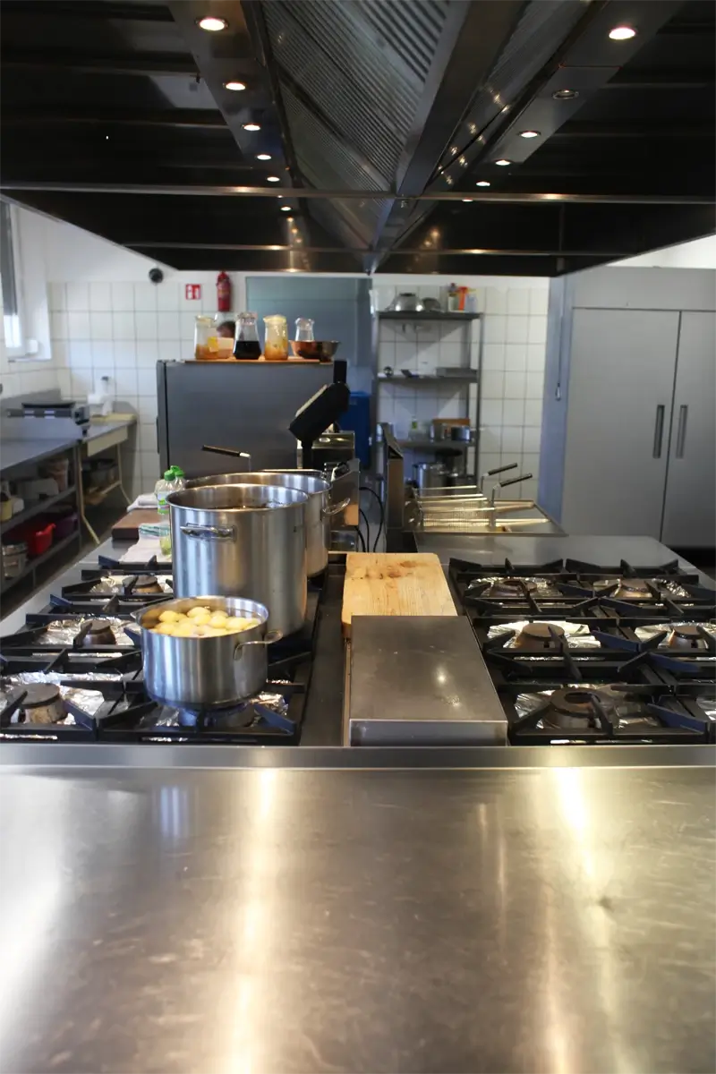 Centralna wyspa grzewcza w kuchni gastronomicznej z kuchnią gazową, frytkownicą, deską do krojenia i okapem sufitowym.