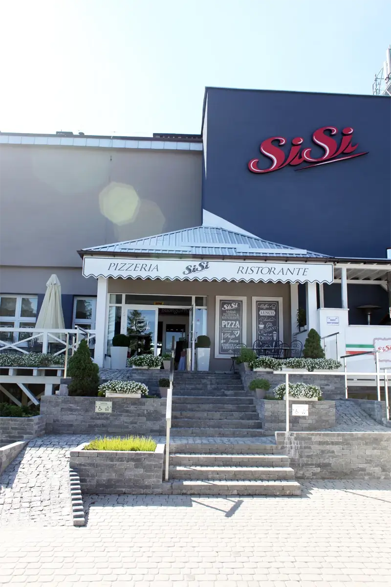 Wejście do restauracji SiSi – pizzeria i ristorante zlokalizowana w nowoczesnym budynku, z zewnętrznymi schodami, białym markizem i dekoracyjną roślinnością.