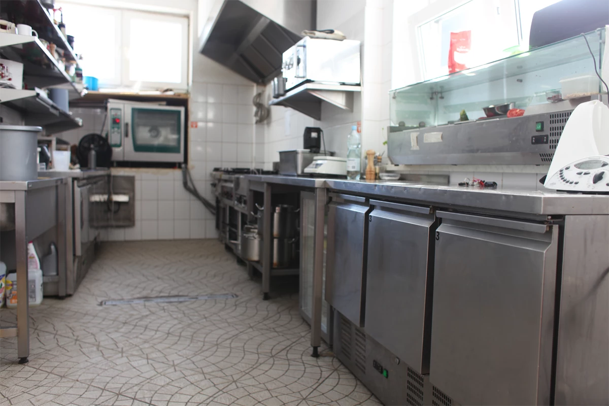 Widok na przestronną, profesjonalną kuchnię gastronomiczną z piecem konwekcyjno-parowym, ladą chłodniczą, stołami roboczymi ze stali nierdzewnej i urządzeniami kuchennymi w tle.