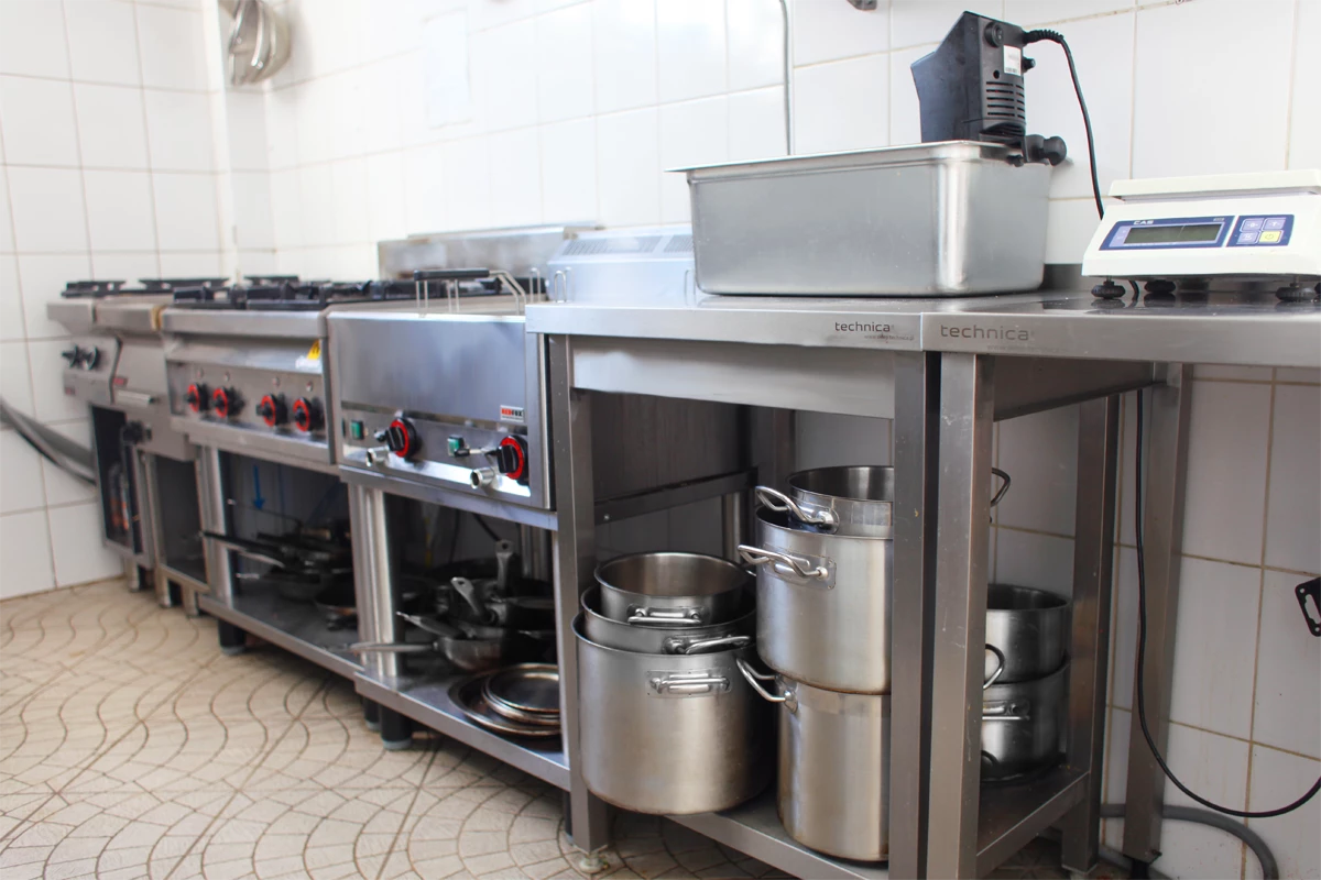 Profesjonalna kuchnia gastronomiczna z linią urządzeń do gotowania, w tym kuchnią gazową, frytownicą oraz stołem roboczym marki Technica, pod którym przechowywane są garnki i naczynia.
