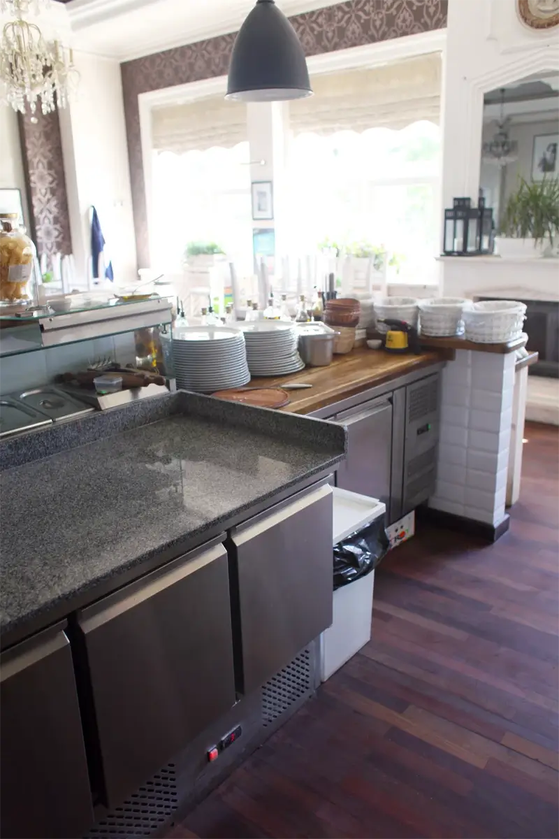 Stanowisko robocze w restauracji z granitowym blatem, chłodnią gastronomiczną podblatową, witryną chłodniczą i uporządkowanymi naczyniami, w eleganckim, jasnym wnętrzu z drewnianą podłogą.