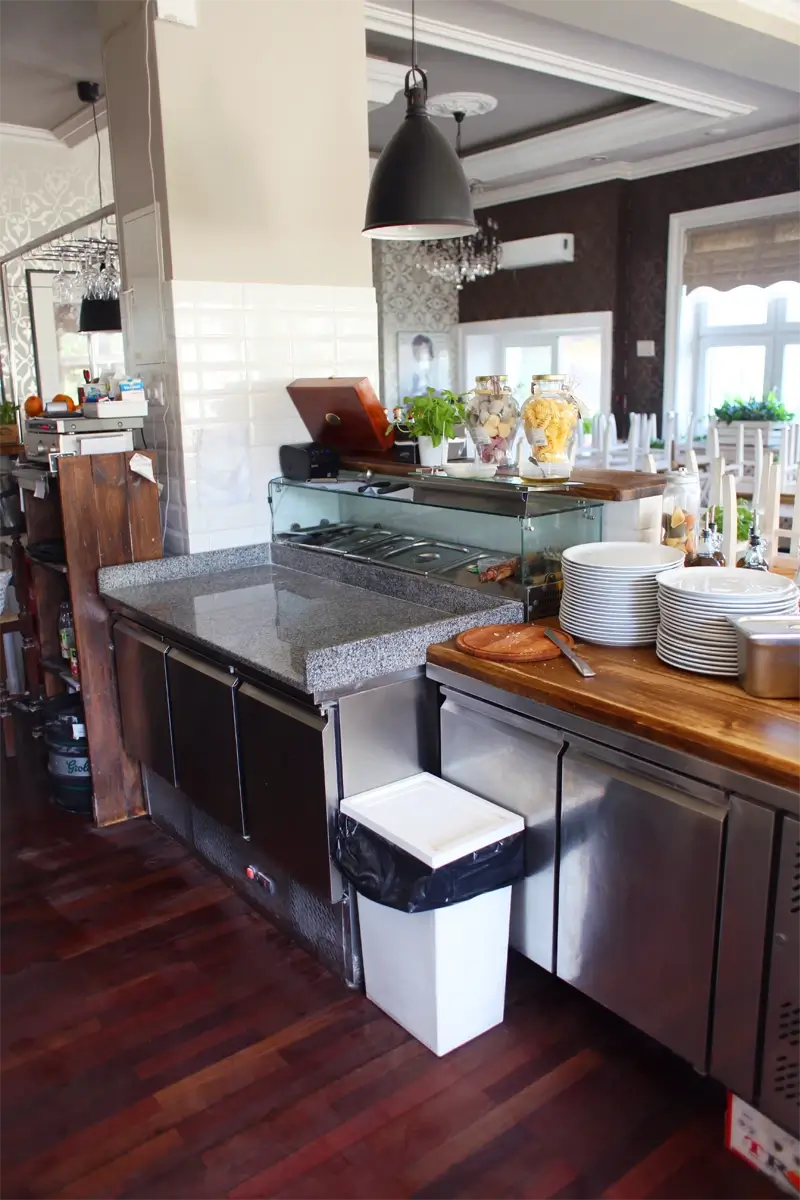 Stanowisko robocze w restauracji z granitowym blatem, chłodnią podblatową i witryną chłodniczą z pojemnikami na składniki, uzupełnione o przygotowane talerze i sztućce — eleganckie wnętrze z drewnianą podłogą.