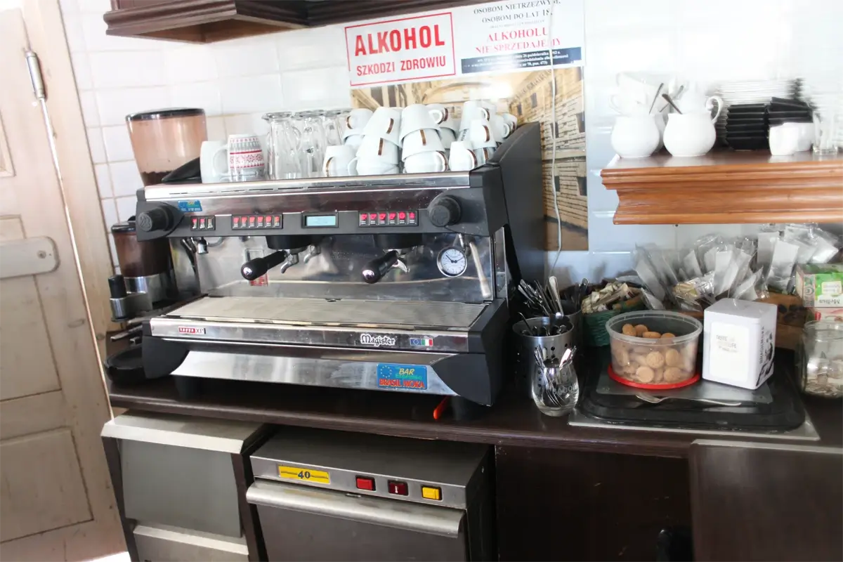 Profesjonalny ekspres ciśnieniowy do kawy w kawiarni, otoczony filiżankami, młynkiem, łyżeczkami, cukrem i dodatkami – kompletne stanowisko baristy z oznaczeniem „Alkohol szkodzi zdrowiu” na tle.