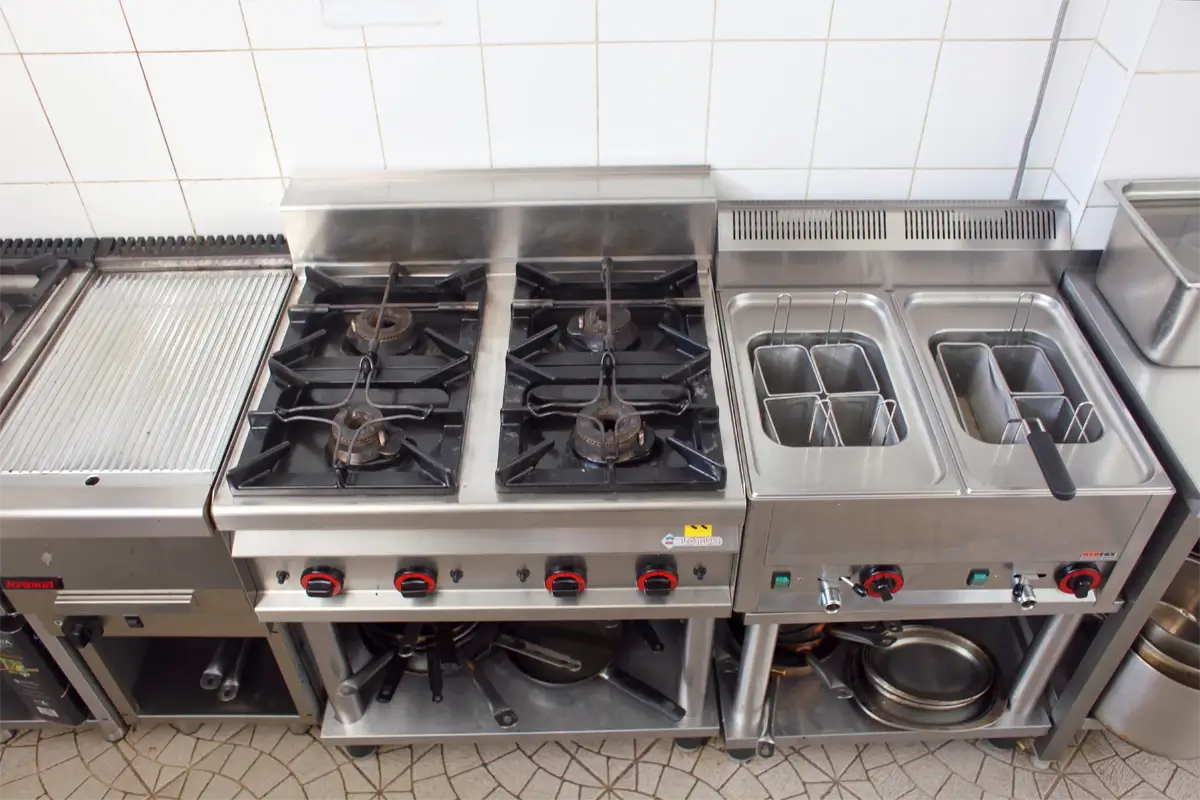 Profesjonalna linia gastronomiczna – kuchenka gazowa czteropalnikowa, frytkownica gastronomiczna z podwójnym zbiornikiem oraz grill żebrowany, wszystko wykonane ze stali nierdzewnej i zainstalowane w kuchni restauracyjnej.