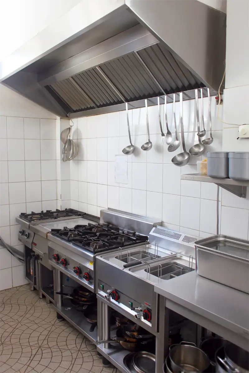 Stanowisko kuchenne w profesjonalnej kuchni gastronomicznej – kuchenki gazowe wielopalnikowe, frytownica, naczynia ze stali nierdzewnej i chochle zawieszone pod wyciągiem kuchennym, ściana wyłożona kafelkami.
