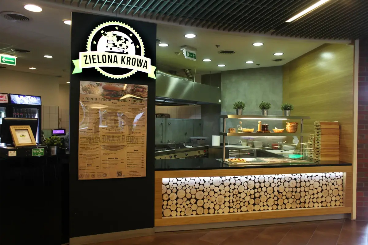 Nowoczesny lokal gastronomiczny Zielona Krowa z otwartą kuchnią, ladą ekspozycyjną i menu na ścianie, znajdujący się w centrum handlowym. Wyróżniający się design z elementami drewna i industrialnym wykończeniem.