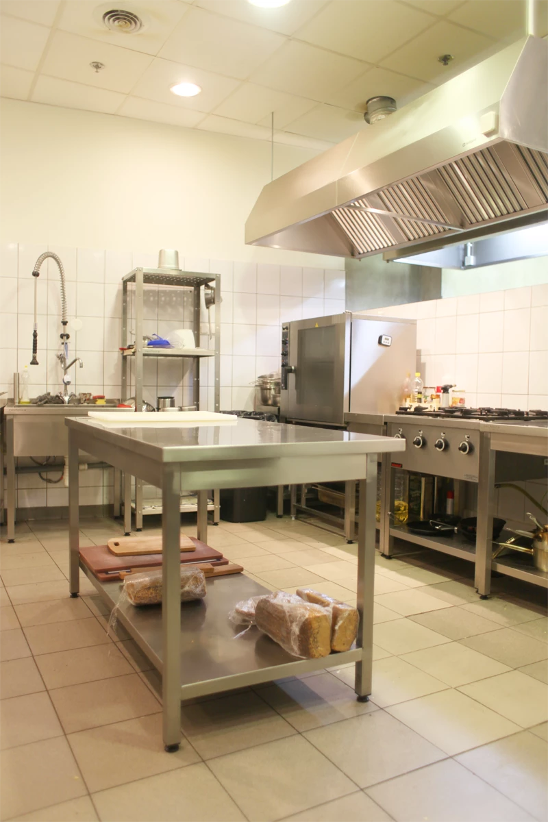 Nowoczesna kuchnia gastronomiczna z centralnym stołem roboczym ze stali nierdzewnej, z półką dolną na pieczywo, stanowiskiem zlewowym, regałem, kuchnią gazową oraz wydajnym okapem kuchennym.