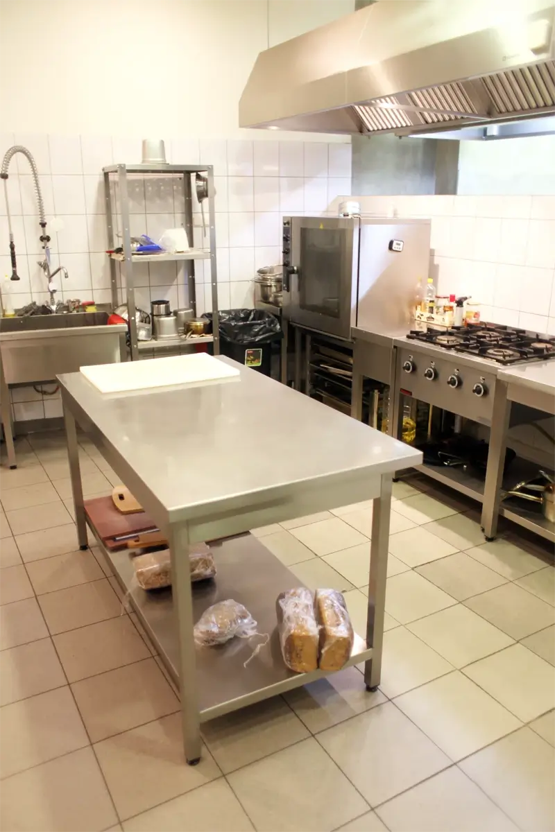 Profesjonalna kuchnia gastronomiczna z centralnym stołem roboczym ze stali nierdzewnej, piekarnikiem konwekcyjno-parowym, kuchnią gazową, okapem i regałem magazynowym, idealna do intensywnej pracy w lokalu gastronomicznym.
