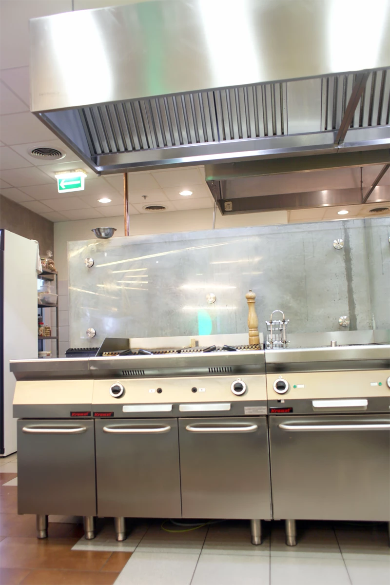 Stanowisko kuchenne z profesjonalnymi urządzeniami gastronomicznymi – kuchnią gazową z grillem i planchą, zlokalizowane pod dużym okapem wentylacyjnym ze stali nierdzewnej, w nowoczesnej kuchni restauracyjnej.