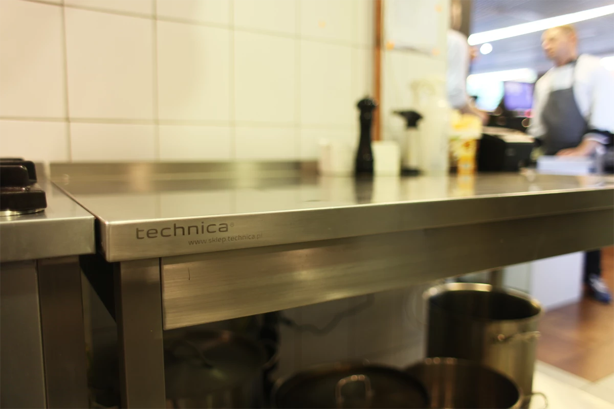 Zbliżenie na stół roboczy ze stali nierdzewnej marki Technica w profesjonalnej kuchni gastronomicznej, z widocznym logo i adresem sklepu internetowego, w tle kucharze przy stanowisku pracy.