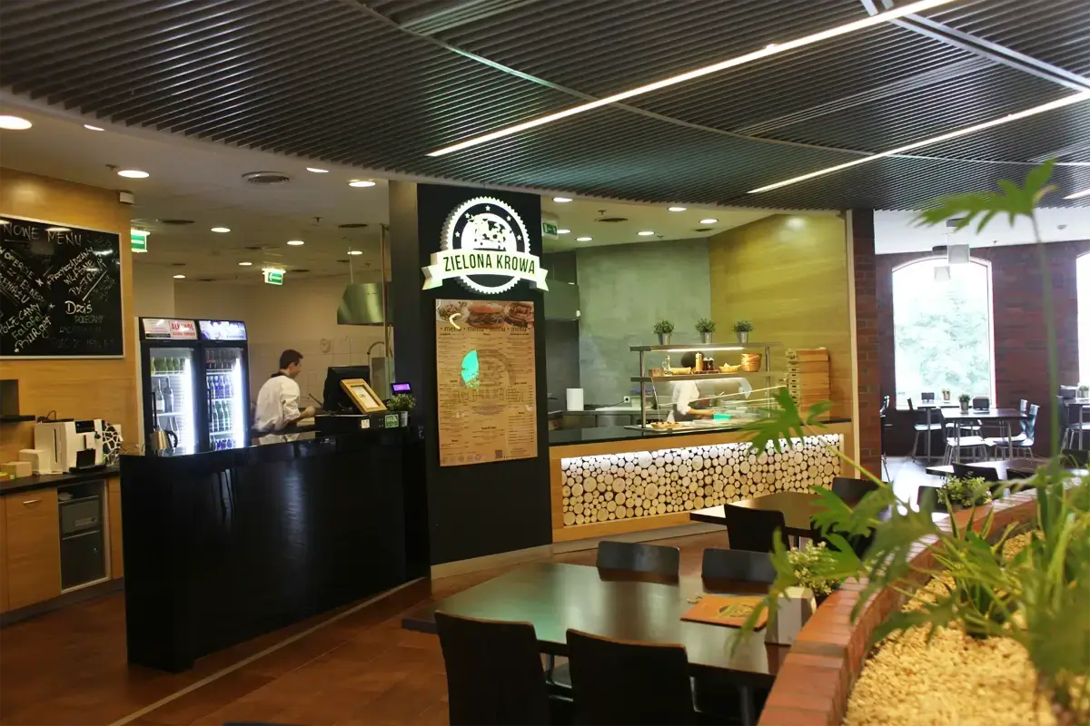 Wnętrze lokalu gastronomicznego Zielona Krowa – widok na strefę zamówień z logo, menu na ścianie, ladę ekspozycyjną oraz stoliki dla gości w przestronnej, nowoczesnej aranżacji z elementami zieleni.