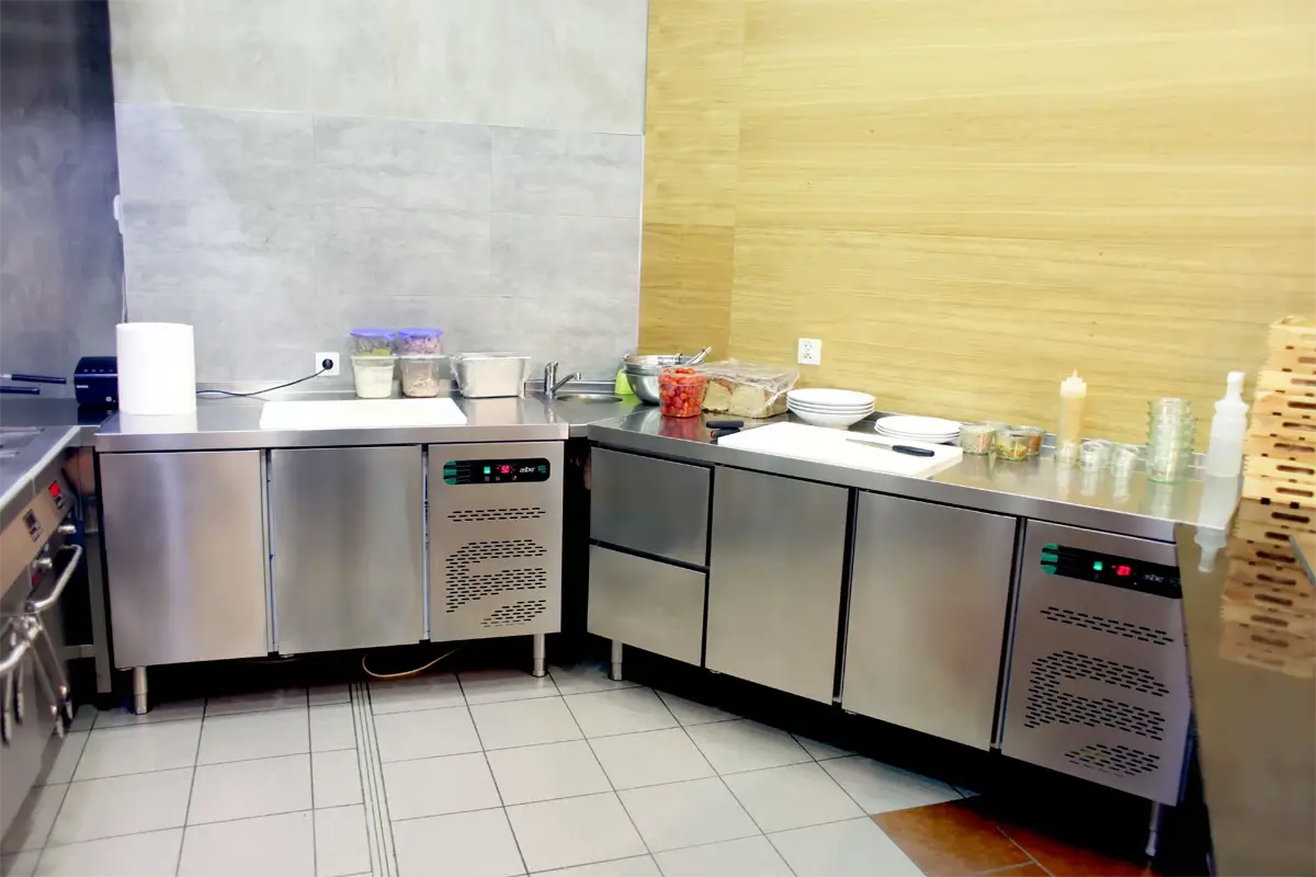 Stanowisko przygotowawcze w kuchni gastronomicznej – zabudowa chłodnicza ze stali nierdzewnej z blatami roboczymi, pojemnikami GN, deską do krojenia, talerzami oraz pojemnikami z warzywami.