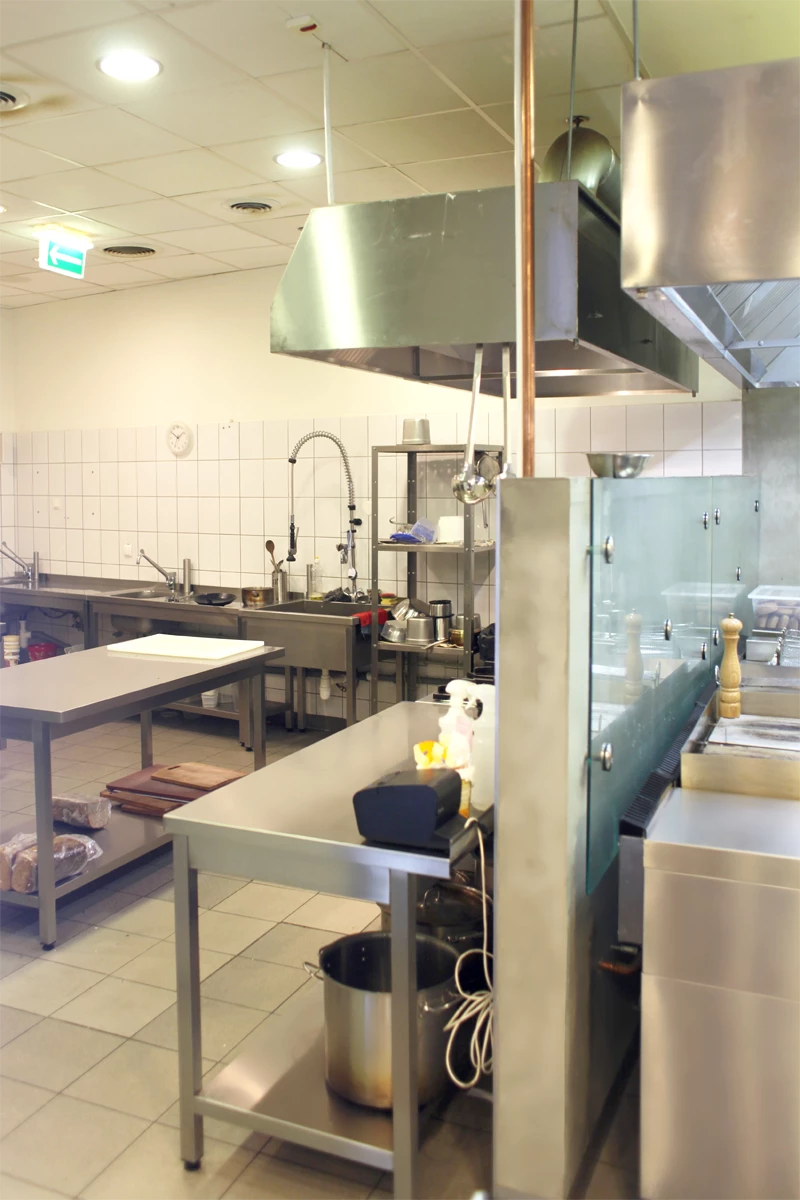 Profesjonalna kuchnia gastronomiczna wyposażona w stoły robocze ze stali nierdzewnej, zlew dwukomorowy, wyciągi wentylacyjne oraz urządzenia do przygotowania potraw, widok ogólny zaplecza kuchennego w restauracji.