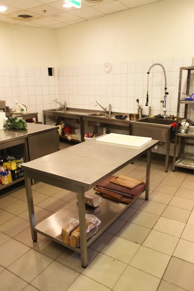 Profesjonalne stanowisko robocze w kuchni gastronomicznej — stół ze stali nierdzewnej z półką dolną, deskami do krojenia, krajalnicą oraz przestrzenią do mycia naczyń i warzyw.