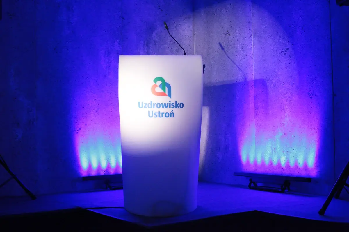 Podświetlana mównica z logo Uzdrowisko Ustroń na tle betonowej ściany z fioletowym oświetleniem – przygotowanie sceny na konferencję uzdrowiskową, prezentację branżową lub wydarzenie z zakresu turystyki medycznej i zdrowia publicznego.