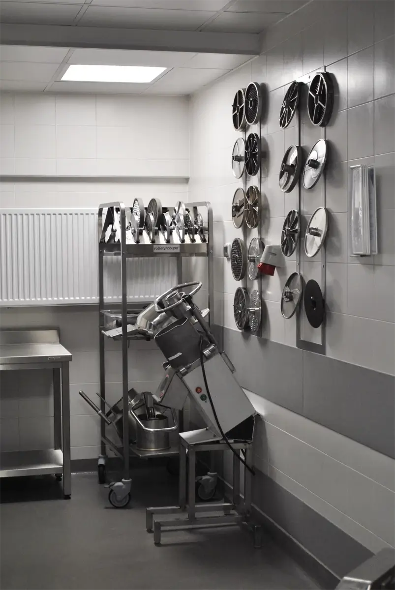 Stanowisko z profesjonalną krajalnicą gastronomiczną do warzyw, marki Robot Coupe, wraz z pełnym zestawem tarcz tnących na regale i ścianie.
