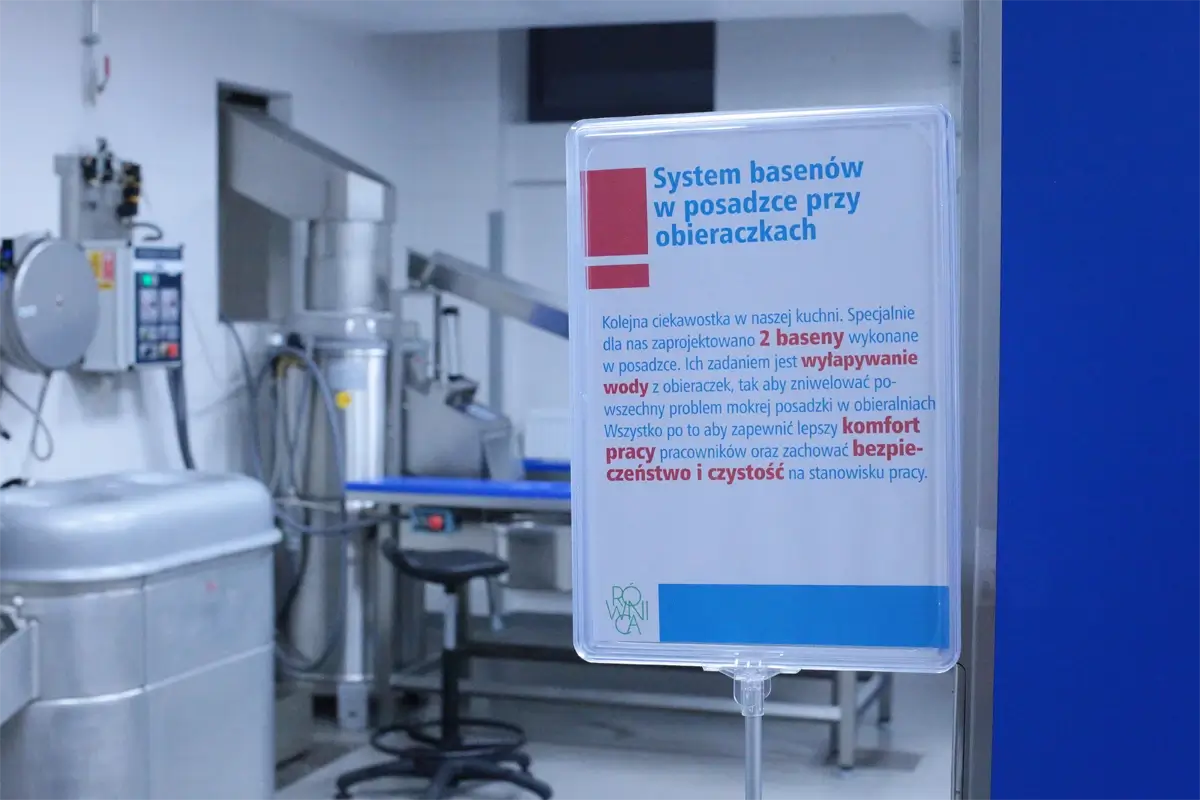 Tablica informacyjna opisująca system basenów w posadzce przy obieraczkach – rozwiązanie technologiczne eliminujące problem wypływania wody z maszyn do obróbki warzyw, zwiększające komfort pracy oraz bezpieczeństwo i higienę w kuchni zbiorowego żywienia.