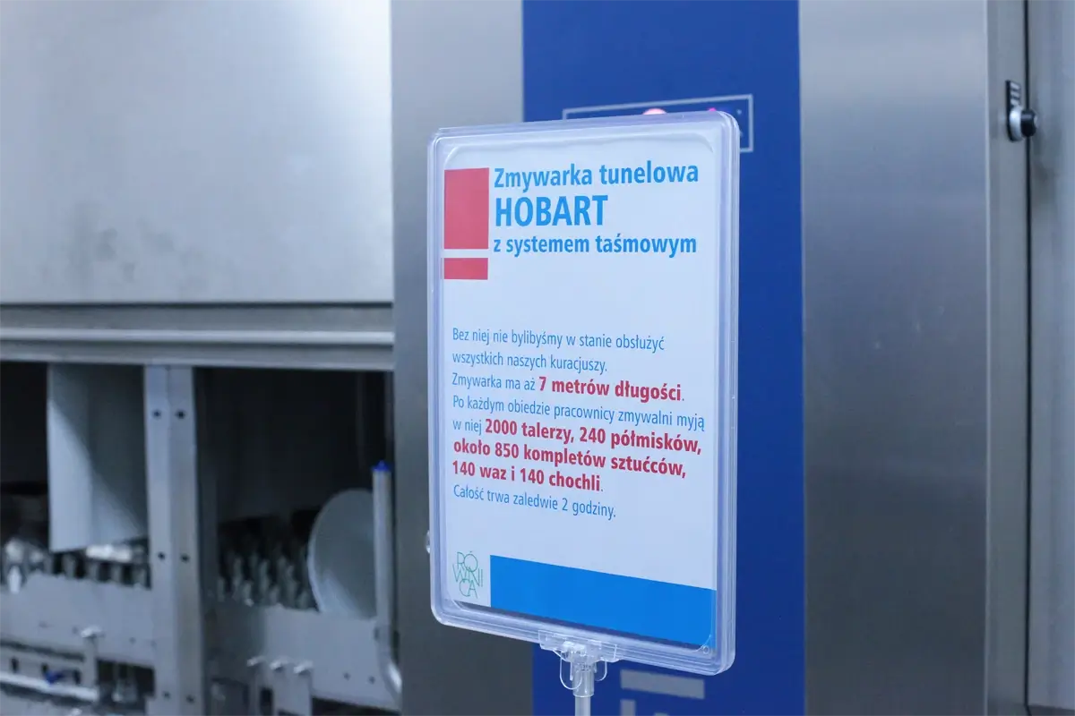 Tabliczka informacyjna przy zmywarce tunelowej Hobart z systemem taśmowym – szczegółowe dane dotyczące długości urządzenia i jego wydajności: mycie 2000 talerzy, 240 półmisków, 850 kompletów sztućców oraz 140 waz i chochli w ciągu 2 godzin.