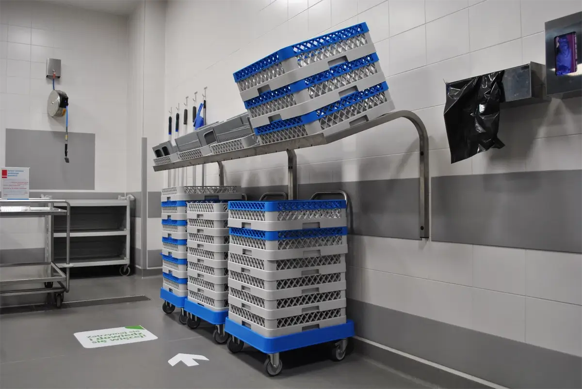Strefa zmywalni w profesjonalnym zapleczu kuchennym – wózki transportowe z plastikowymi koszami zmywarkowymi ustawione pod stalowym regałem ściennym; przestrzeń zaprojektowana z myślą o ergonomii pracy i higienie w gastronomii zbiorowej.
