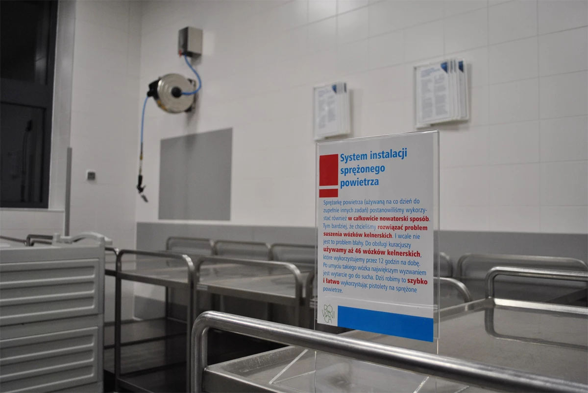Strefa zmywalni w profesjonalnym zapleczu kuchni zbiorowego żywienia – tabliczka informacyjna opisująca system instalacji sprężonego powietrza do suszenia wózków kelnerskich.