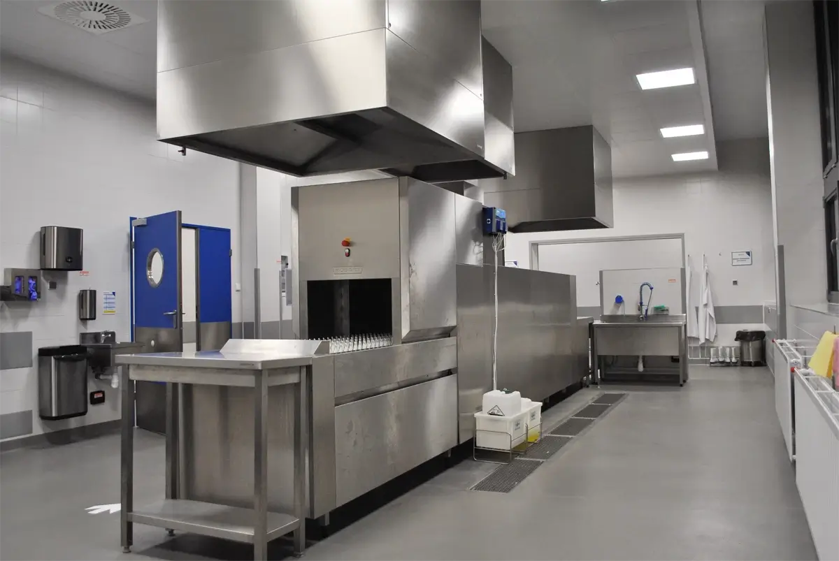Profesjonalna zmywalnia gastronomiczna wyposażona w przemysłową zmywarkę tunelową z systemem okapowym – przestrzeń zaprojektowana zgodnie z normami HACCP, z wydzielonymi strefami do zmywania, suszenia i segregacji naczyń; widoczne środki chemiczne, stół roboczy i komora zlewowa.