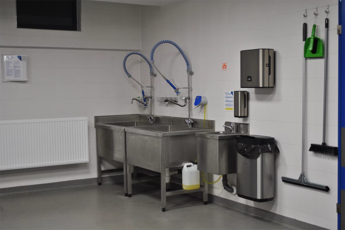 Strefa sanitarna w profesjonalnym zapleczu gastronomicznym – zlew dwukomorowy ze stali nierdzewnej, z elastycznymi wylewkami natryskowymi, osobny zlew do mycia rąk, pojemniki na ręczniki papierowe i środki higieny oraz zestaw do sprzątania naściennego; przestrzeń zgodna z normami HACCP.