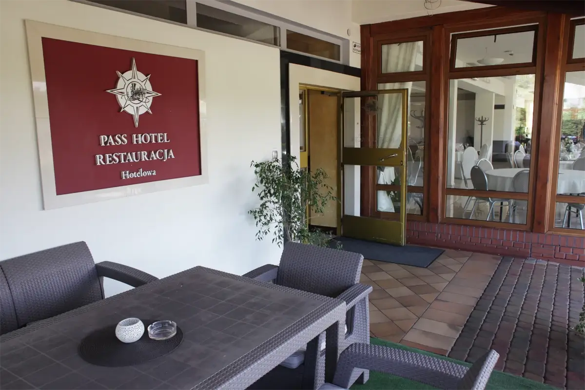 Wejście do restauracji hotelowej Pass Hotel, z widocznym szyldem na ścianie, stolikiem i krzesłami na tarasie oraz otwartymi drzwiami prowadzącymi do wnętrza z nakrytymi stołami.
